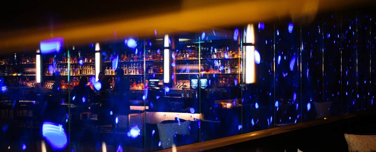 Dimly lit bar with neon blue lights reflecting, showcasing shelves of bottles and cozy seating areas in the background.