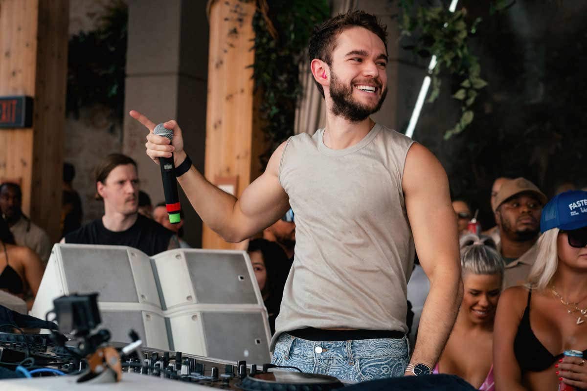 Zedd in the DJ booth at TAO Beach, holding a microphone and smiling, with a crowd of people behind him