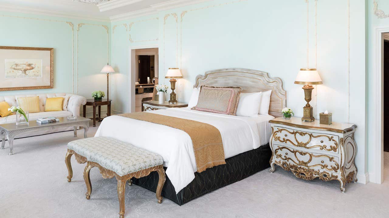 Elegant bedroom with a large bed, ornate furniture, soft blue walls, and warm lighting creating a luxurious ambiance.