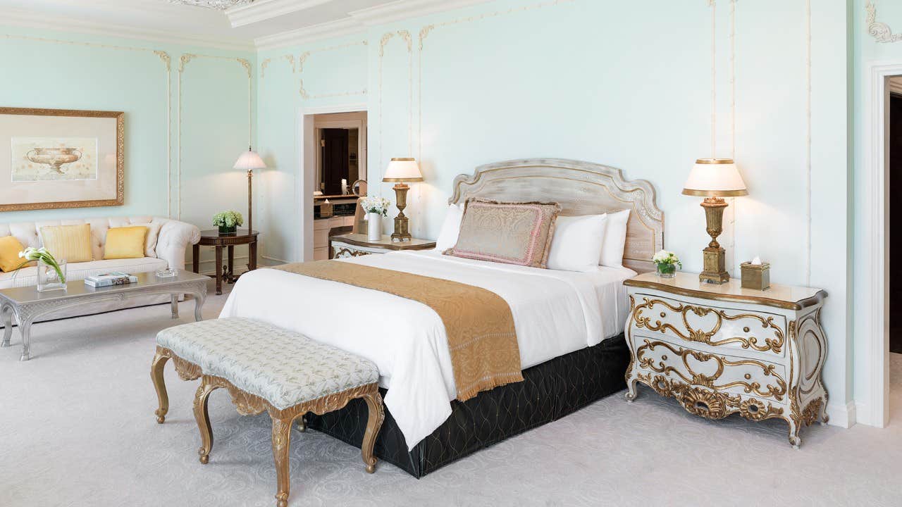 Elegant bedroom with a large bed, ornate furniture, soft blue walls, and warm lighting creating a luxurious ambiance.