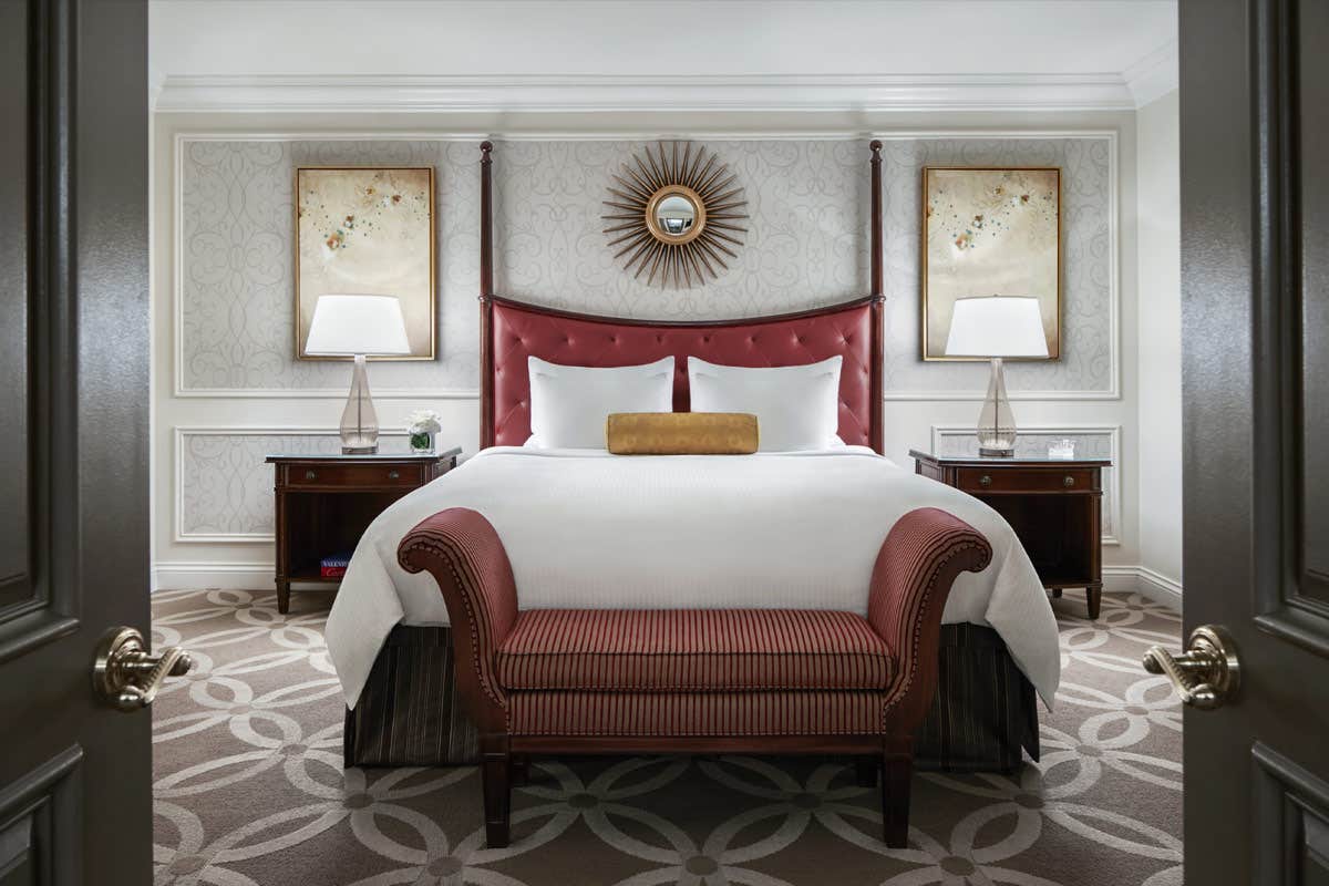 Elegant hotel room with a large bed, red upholstered headboard and bench, bedside tables with lamps, and decorative wall art.