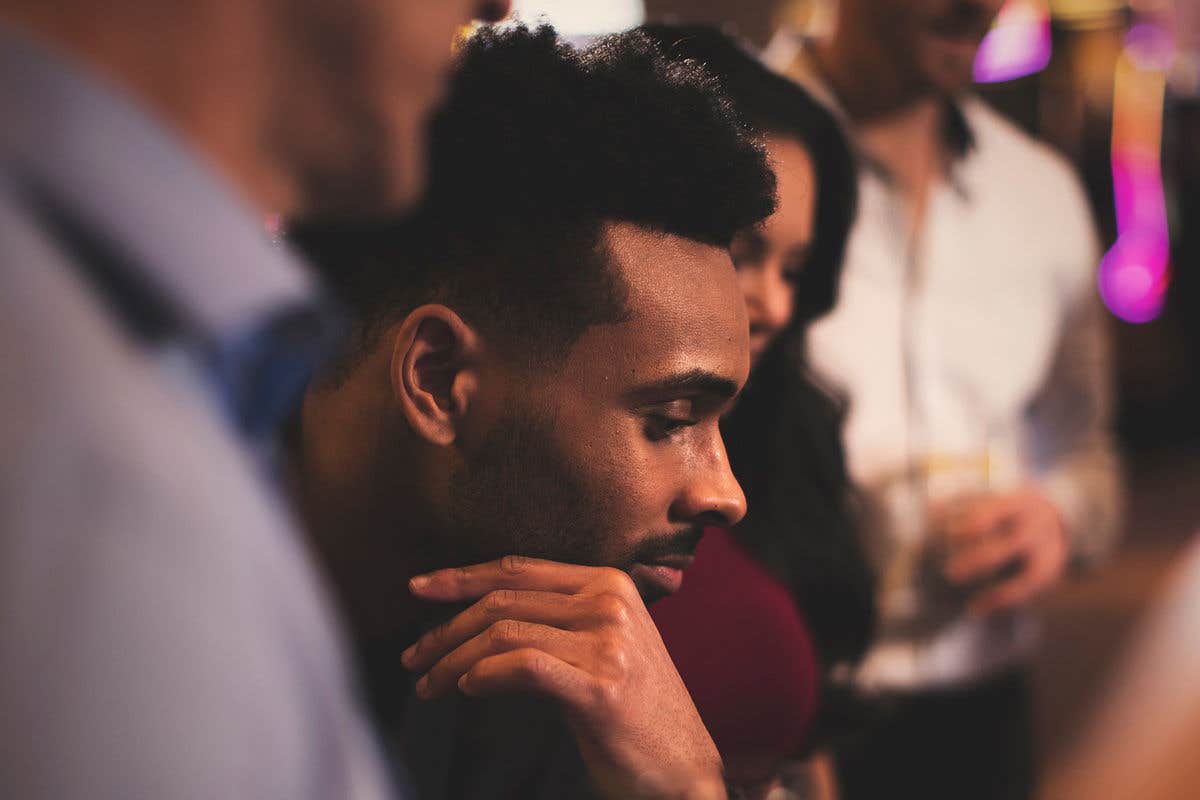 A man with short hair looks thoughtfully downwards; blurred people and lights are in the background.