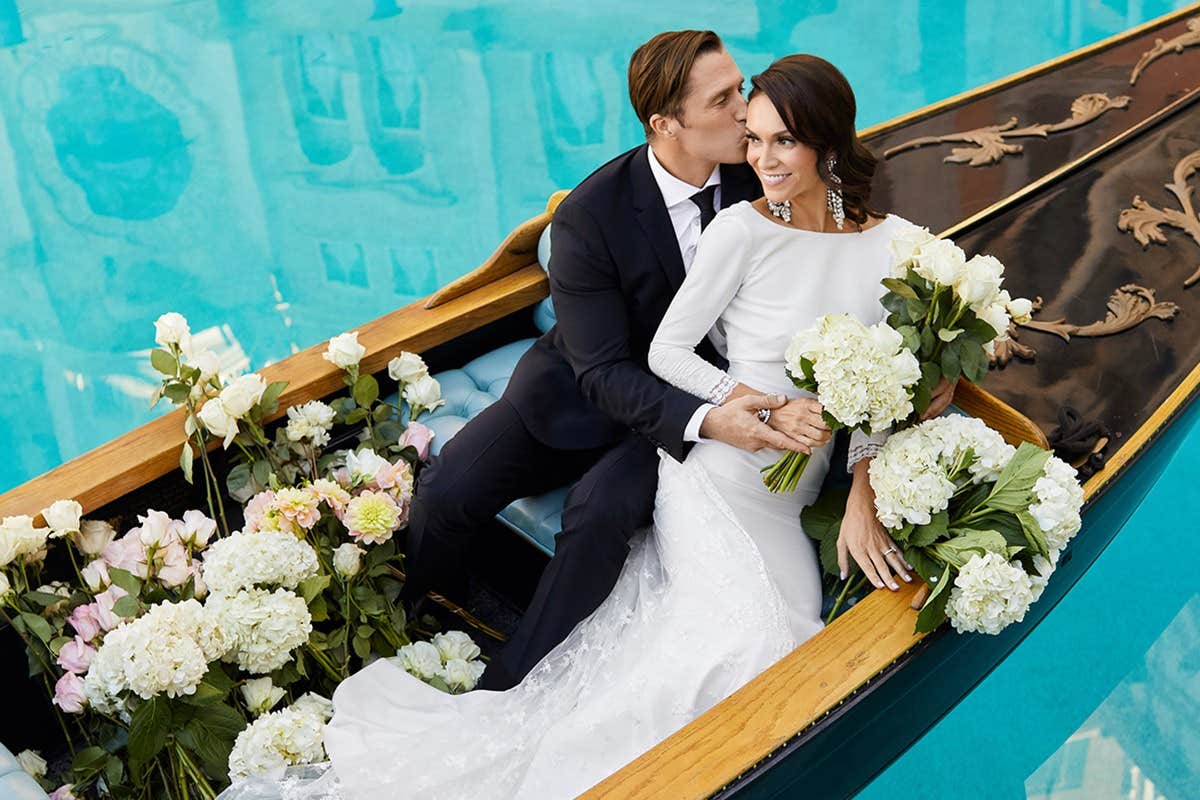 A couple in wedding attire sits in a flower-filled boat; the man kisses the woman's forehead.