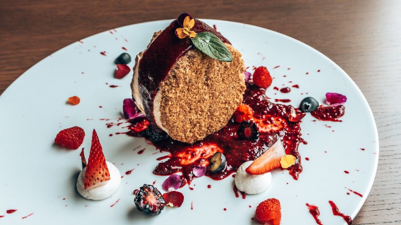 A gourmet dessert on a white plate, topped with berries, cream, and a decorative flower garnish.