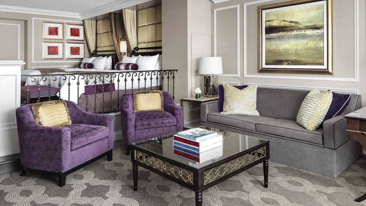 A luxurious living area features purple armchairs, a gray sofa, a glass coffee table with books, and a bedroom in the background.