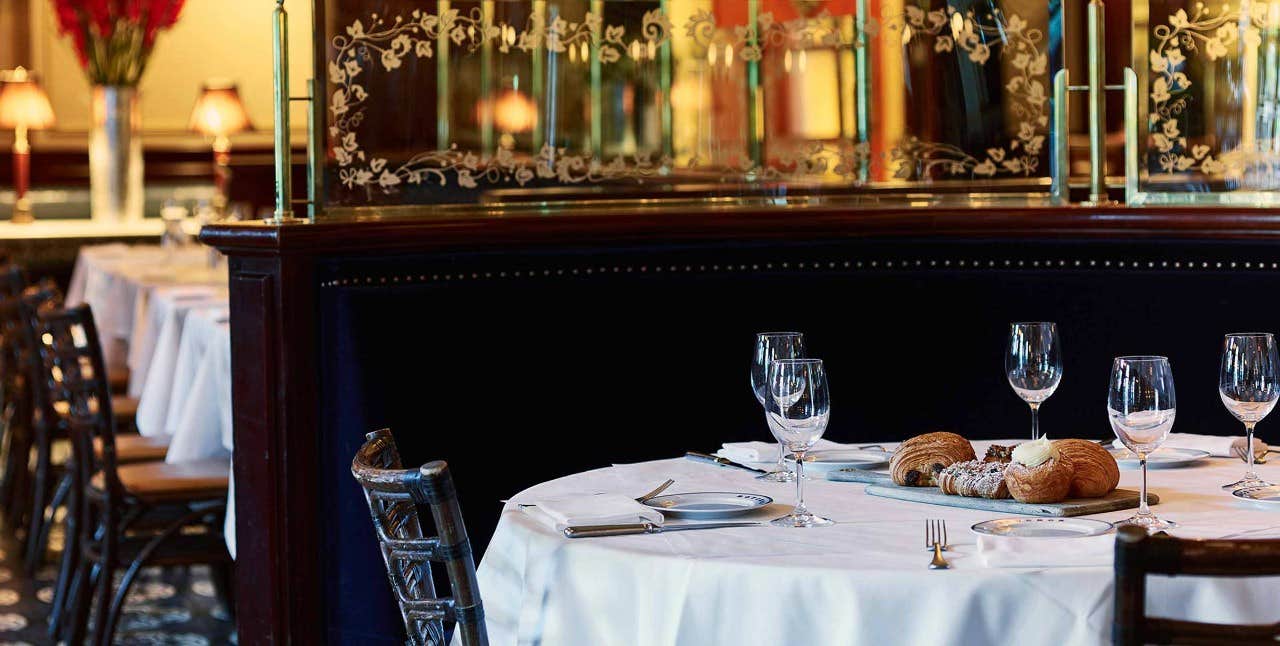 Elegant restaurant with white tablecloths, place settings, wine glasses, and bread on tables, with ornate decor in the background.