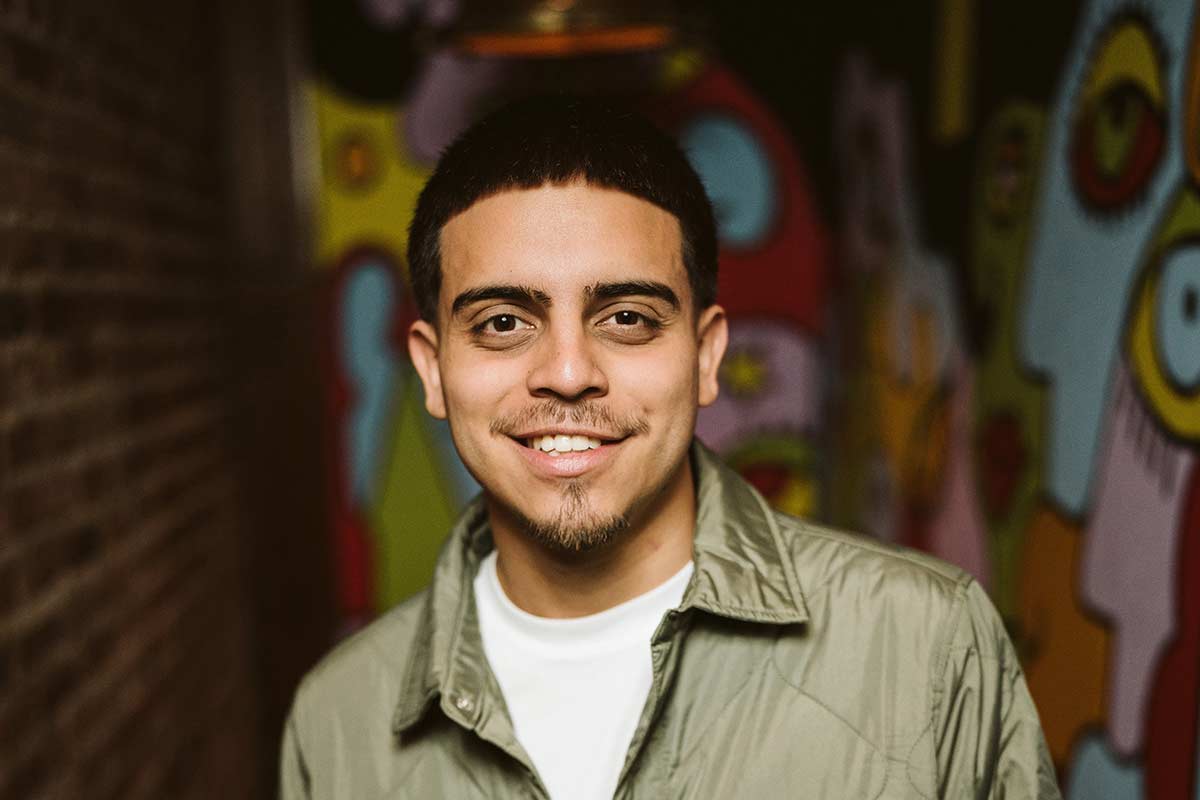 A smiling man in a beige jacket stands against a colorful graffiti wall background.