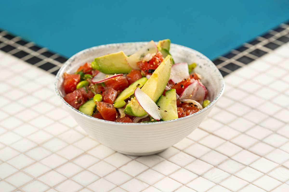 A bowl of fresh poke with tuna, avocado, radish slices, and green onions placed on a tiled surface near a pool.