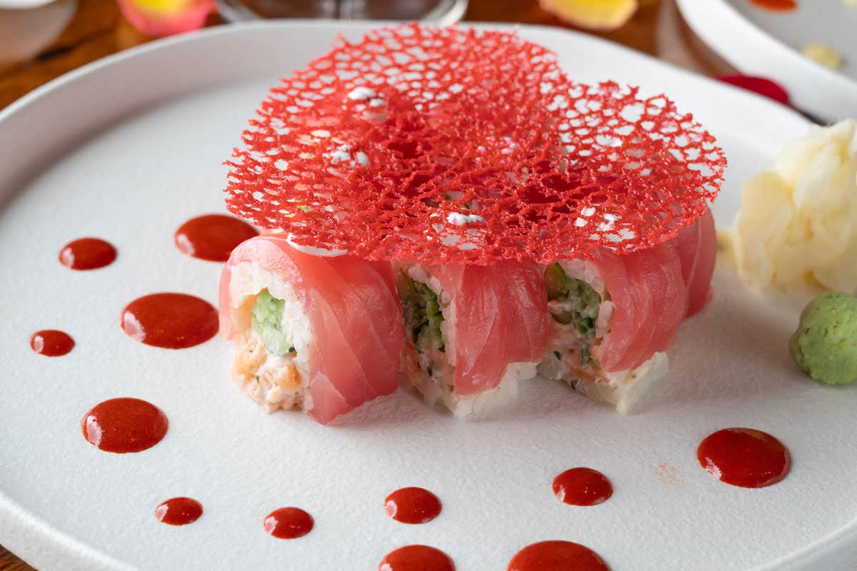 Decorative sushi roll topped with pink tuna and a red lattice, surrounded by red sauce, wasabi, and pickled ginger on a white plate.