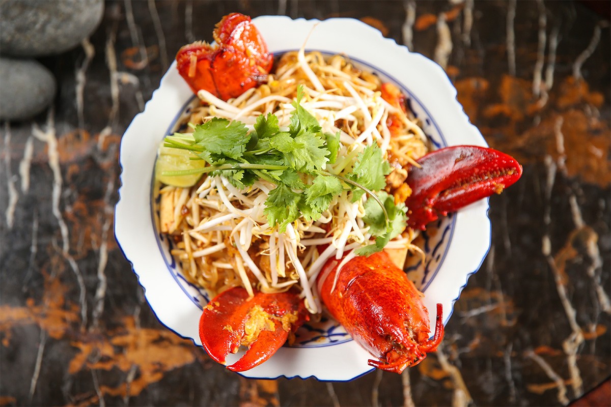 A plate of noodle stir-fry topped with lobster, bean sprouts, and cilantro, served on a dark marbled surface.