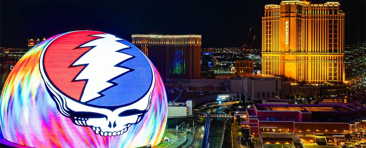 A vibrant, lit-up skyline featuring a glowing dome with a lightning bolt skull design, and illuminated tall buildings at night.