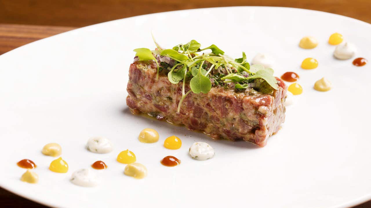 A gourmet dish featuring steak tartare topped with microgreens, surrounded by colorful sauce dollops on a white plate.