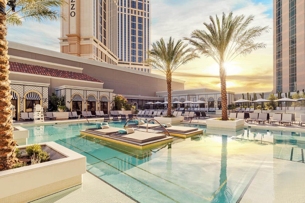 Luxury hotel pool area with lounge chairs, palm trees, and cabanas at sunset, with tall buildings in the background.