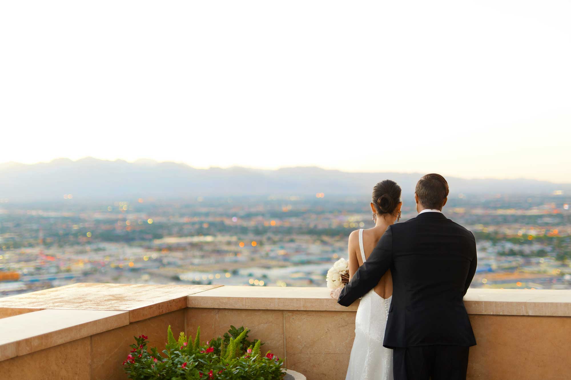 A couple in wedding attire poses on a rooftop with a city skyline in the background