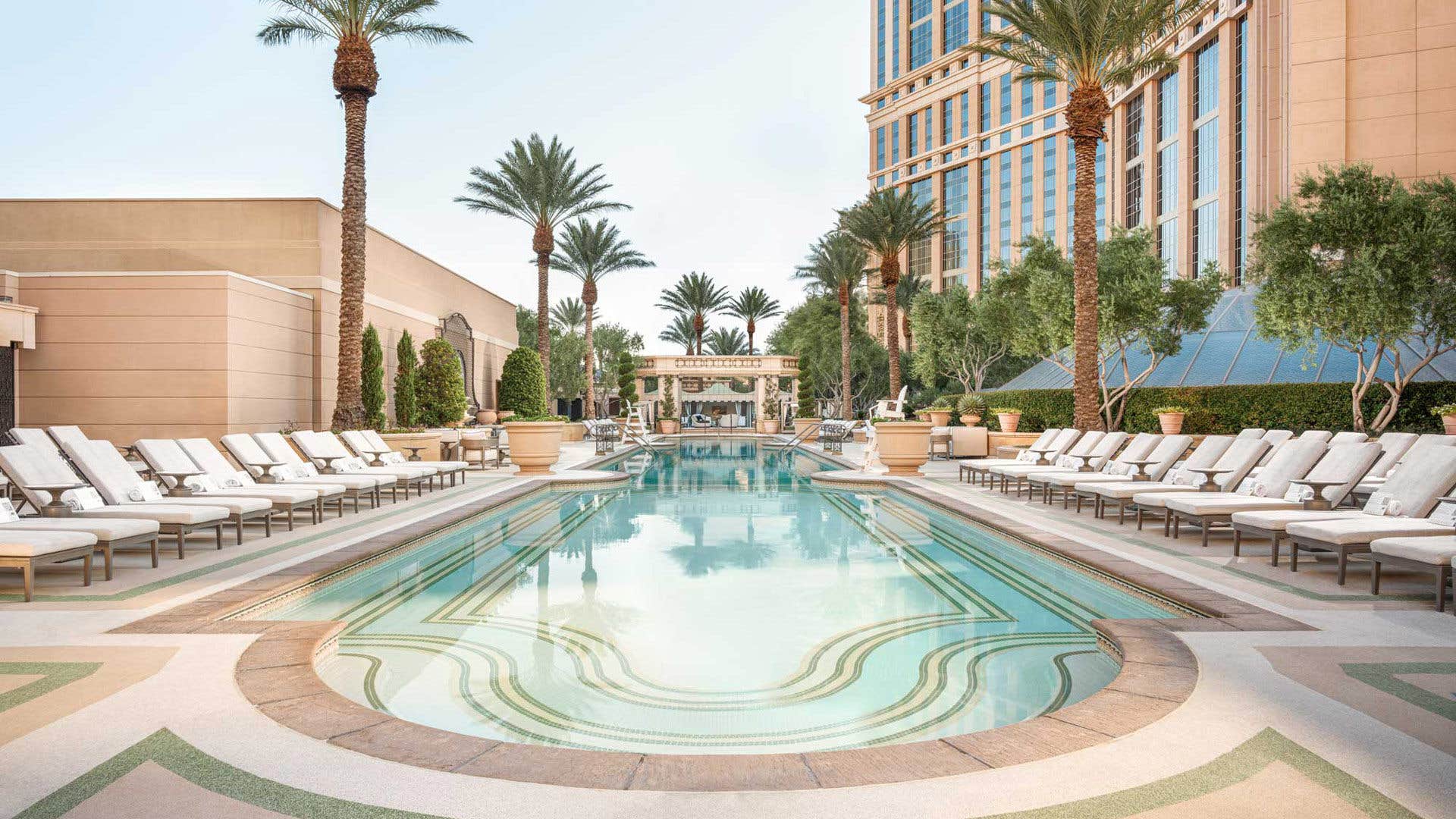 A luxurious swimming pool with lounge chairs, palm trees, and a lavish resort building in the background on a sunny day.