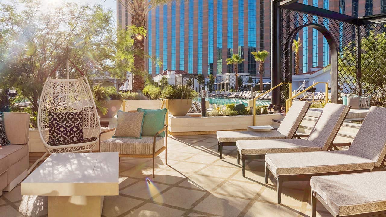 Sunny poolside lounge area with cushioned chairs, wicker chairs, and potted plants; backdrop of modern building and pool.