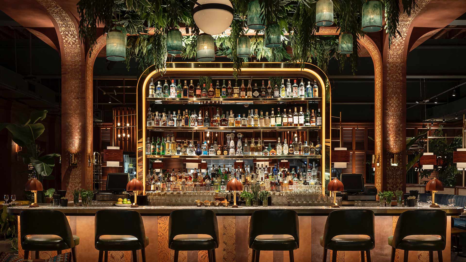 A stylishly decorated bar with a large, illuminated liquor shelf, green barstools, and hanging plants.