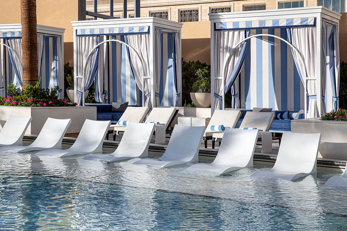 Blue and white poolside cabanas with striped curtains, lounge chairs partially submerged in water.