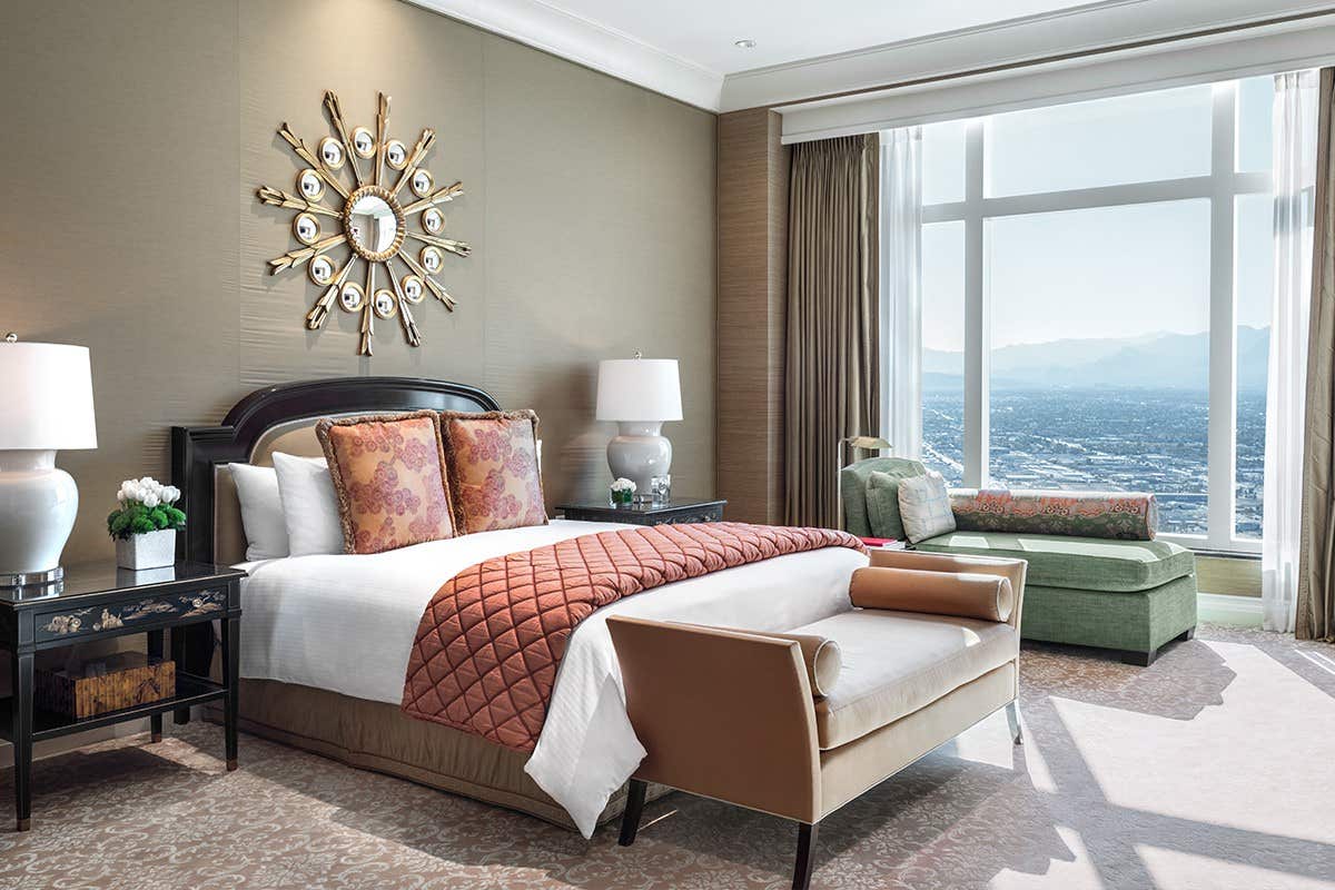 Luxurious hotel room with a large bed, stylish decor, sunburst mirror, and floor-to-ceiling window overlooking a cityscape.