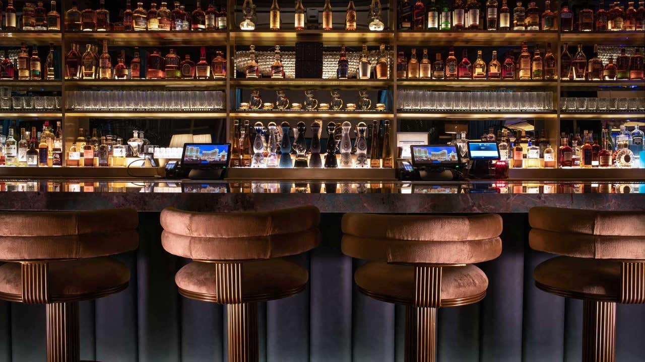 A dimly lit bar with cushioned stools, a variety of liquor bottles, and bar equipment on the shelves.