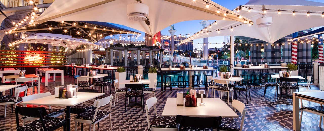 Outdoor restaurant seating area with white tables, string lights, and patterned flooring under a canopy structure.