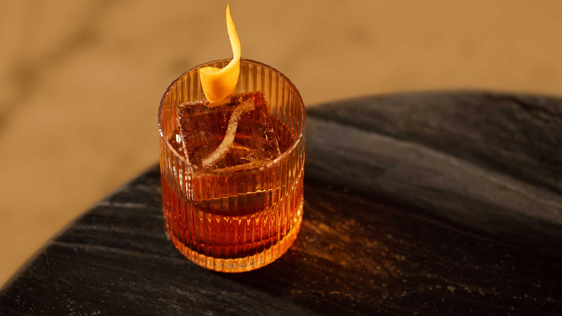 Glass of cocktail with an ice cube and a twist of citrus peel, placed on a dark wooden surface.