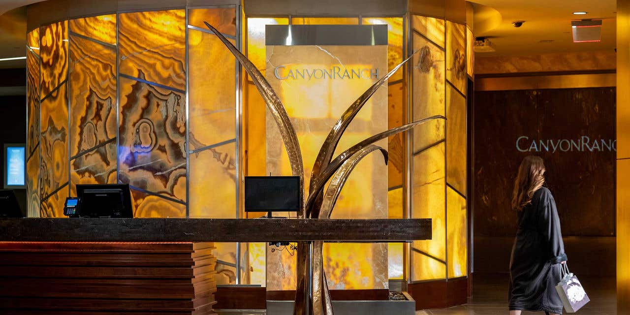 Canyon Ranch Spa and Fitness lobby with a check-in desk to the left and a woman in a robe walking to the treatment rooms on the right.