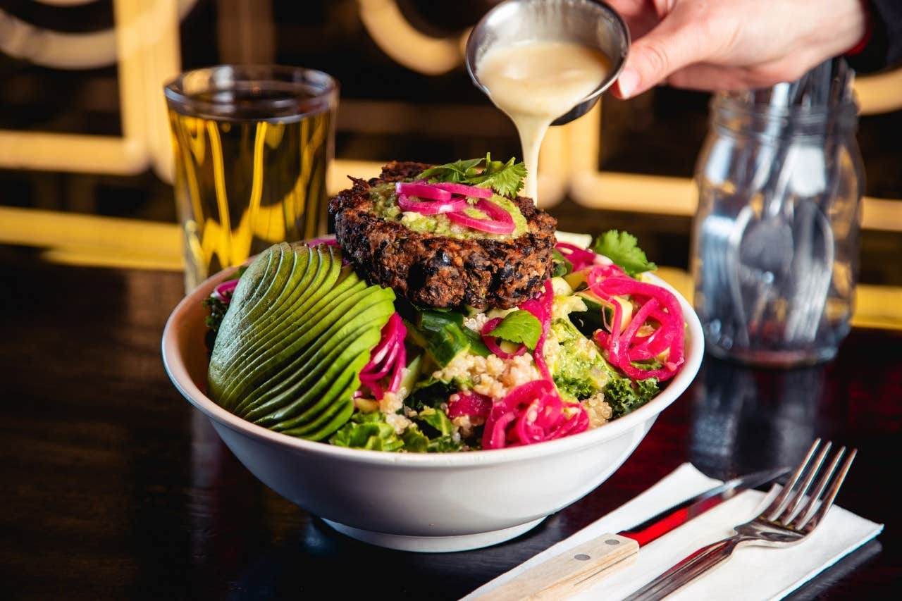 A hand pouring dressing on a bowl of salad topped with a vegetable patty, avocado slices, and pickled onions.