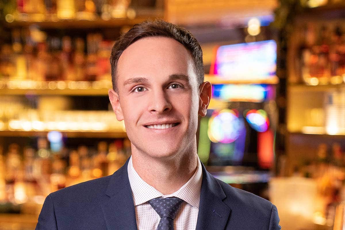 A man in a suit and tie smiles in a warmly lit room with blurred colorful lights in the background.