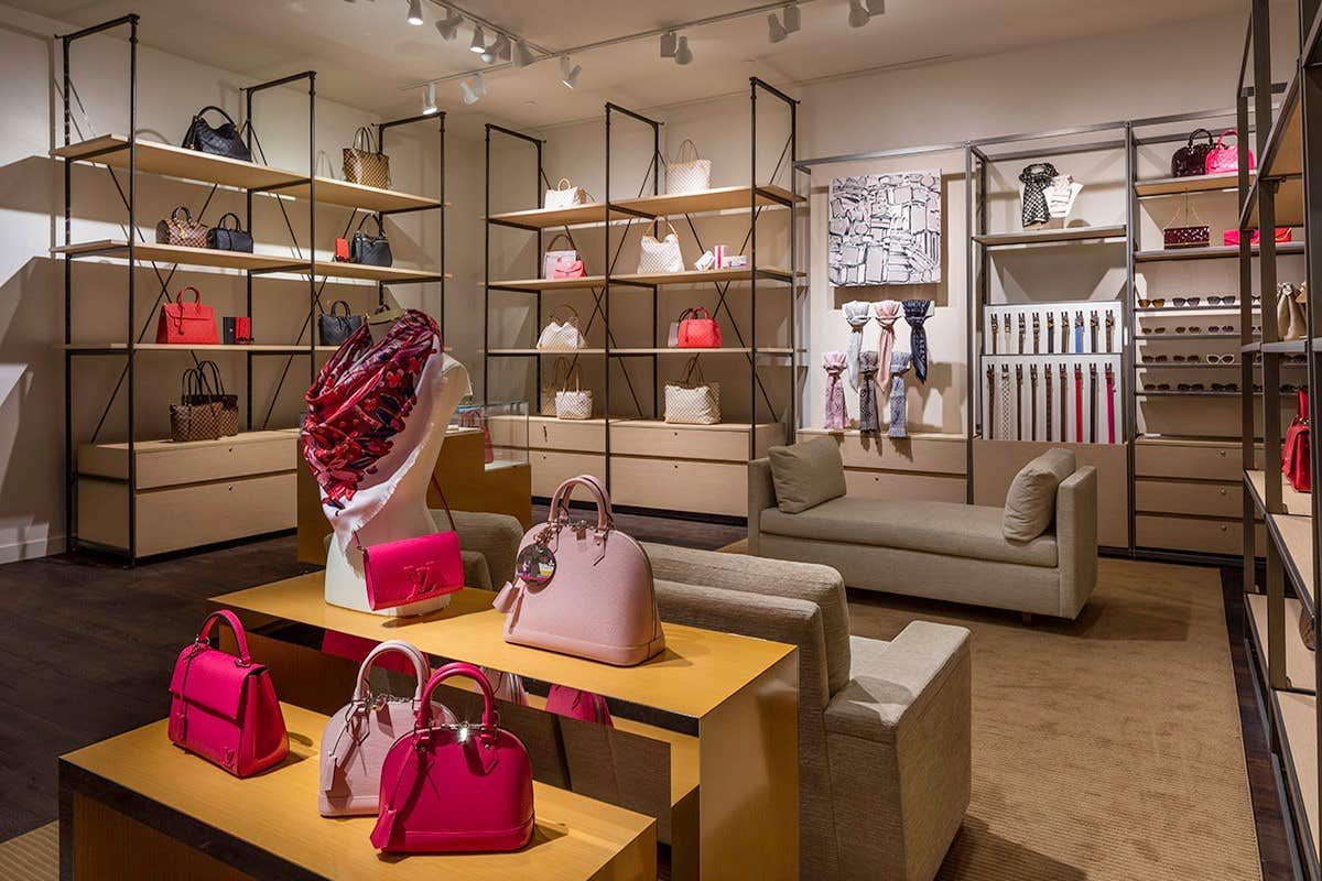 A luxury handbag store with handbags on display shelves and tables, and a couple of chairs for customers to sit.