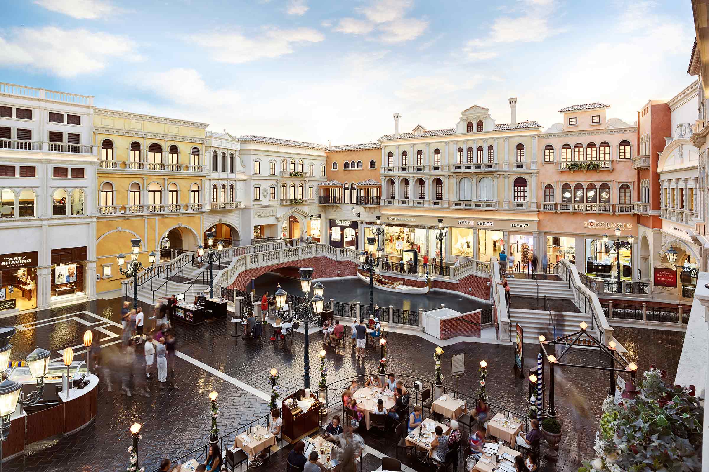 Indoor mall with Venetian-style architecture, featuring a canal, decorative bridge, shops, and a dining area with patrons.