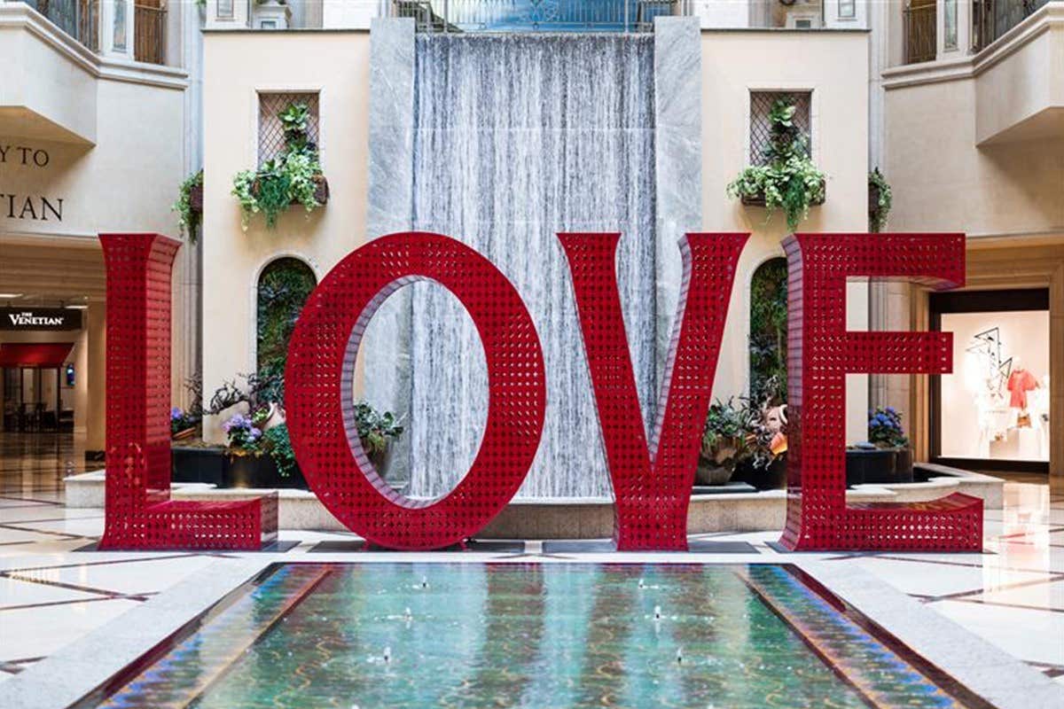 Large red "LOVE" sculpture in front of a cascading waterfall inside a shopping mall, near a clear water fountain.