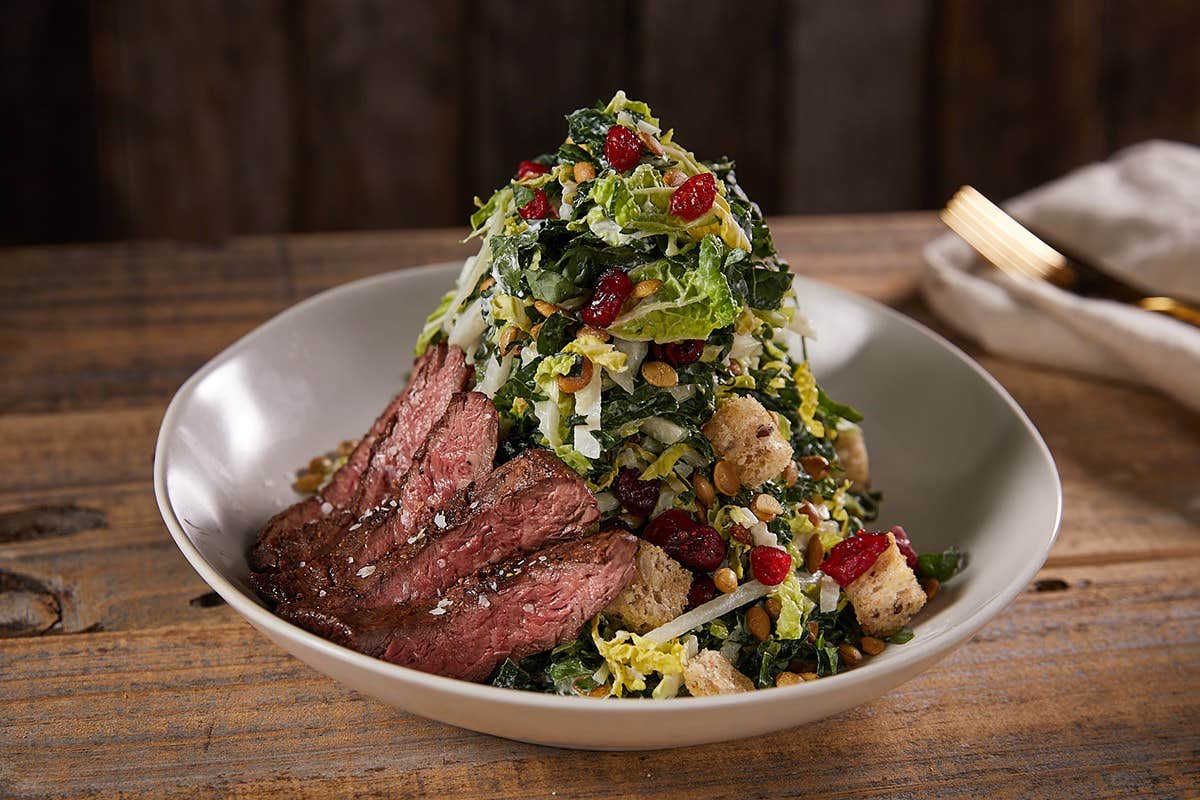 A bowl of mixed green salad topped with grilled steak slices, cranberries, croutons, seeds, and shredded cheese.