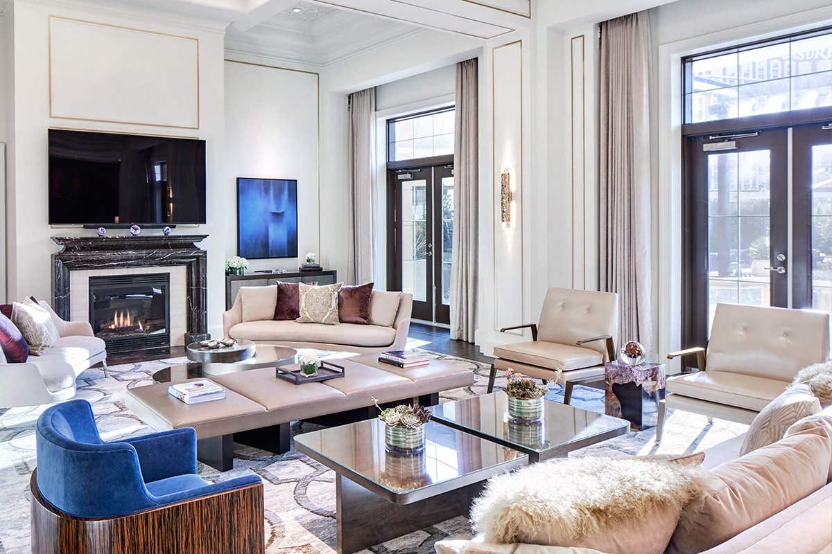 Modern living room with light-colored furniture, a large TV above a fireplace, and large windows letting in natural light.