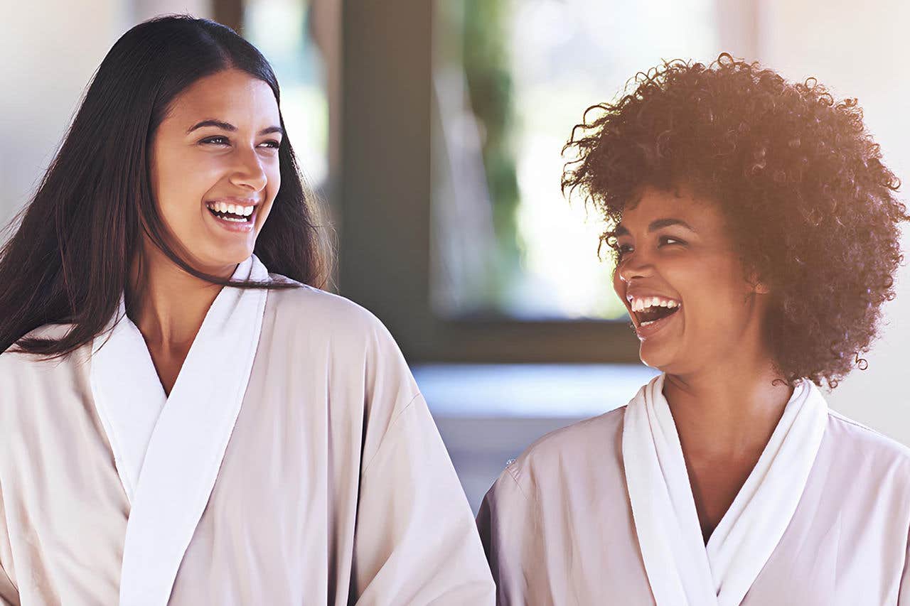 Two women wearing robes laugh together in a bright room, creating a joyful and relaxed atmosphere.