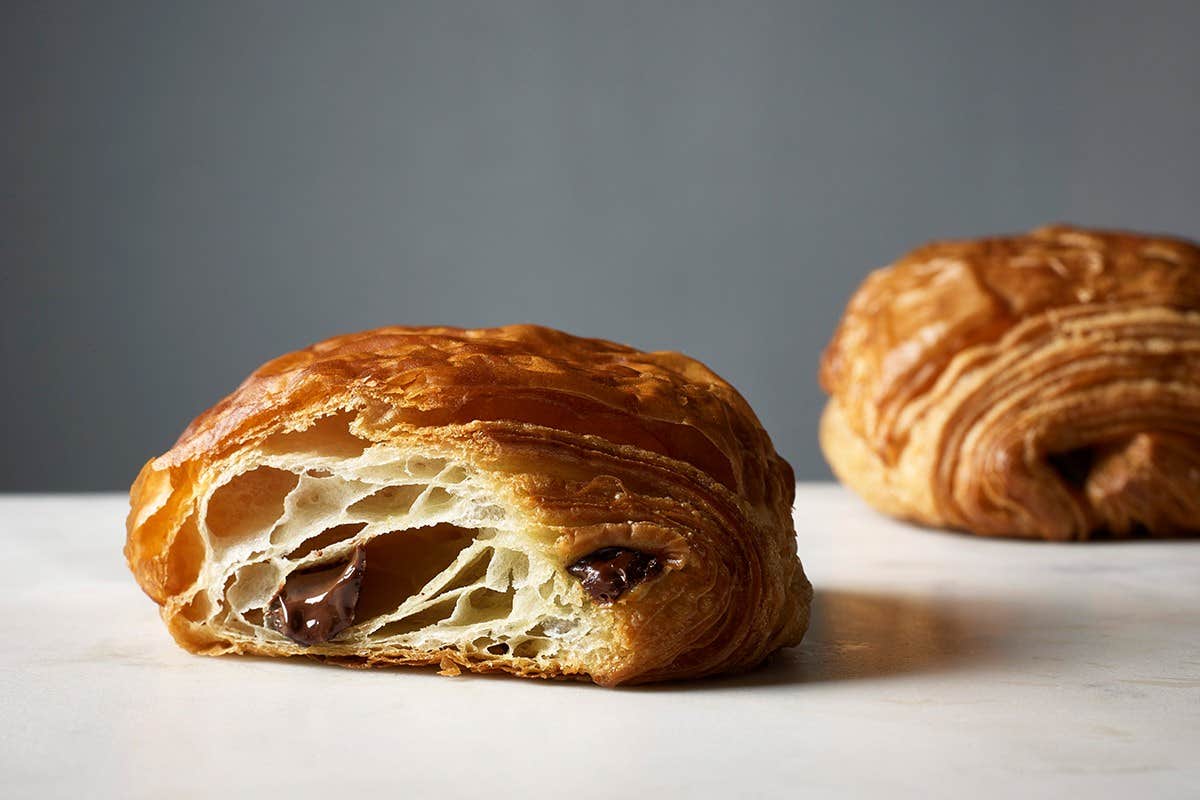 A close-up of a chocolate croissant with another croissant in the background on a light surface.