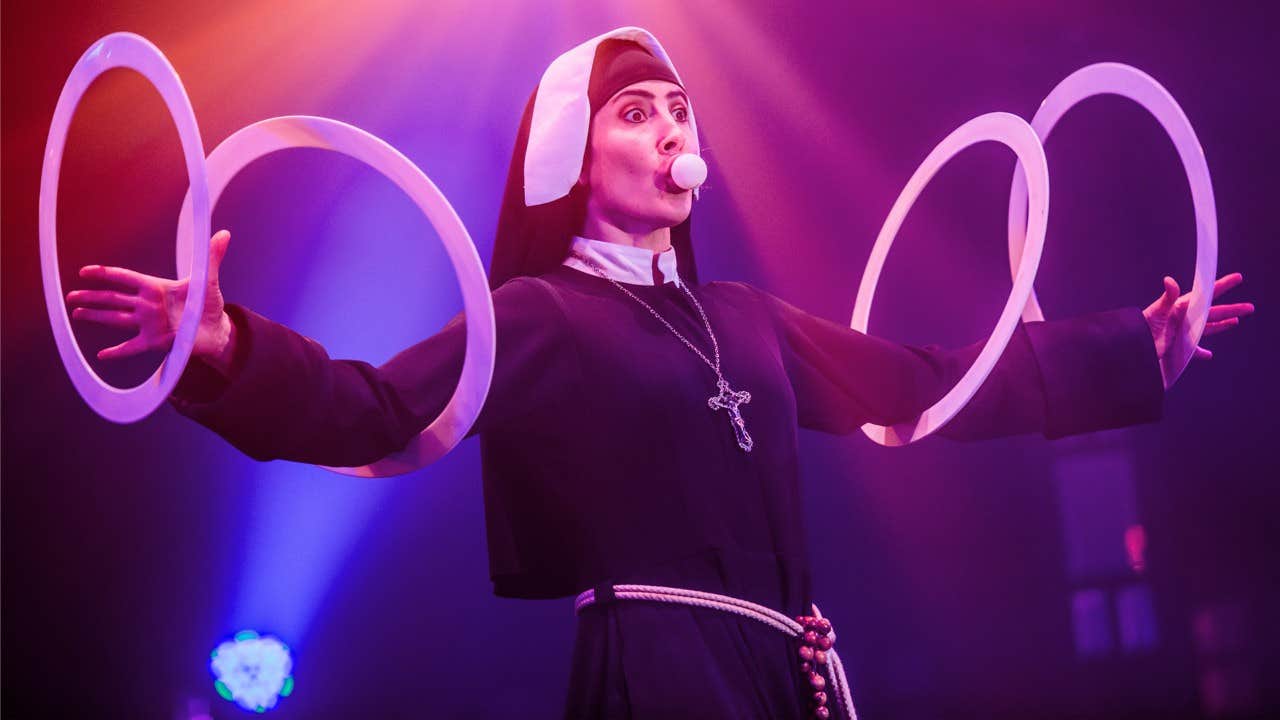 A performer in a nun costume expertly juggles rings under colorful stage lights with a focused expression.