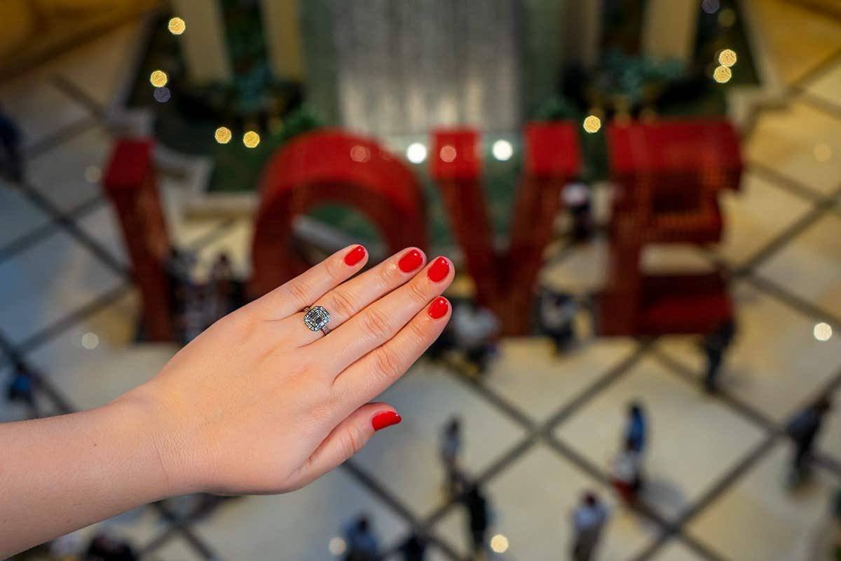 Hand with red nail polish and an engagement ring, in front of large "LOVE" letters and a waterfall in a busy area.