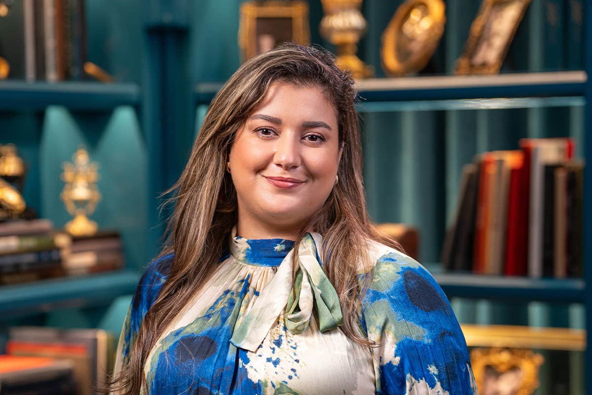 A woman with long hair smiling, wearing a blue and white outfit, standing in front of a bookshelf with framed pictures.