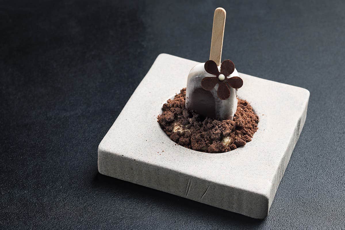 A chocolate ice cream bar with a decorative flower on a stick, presented on a plate with crumbled chocolate and cookie bits.