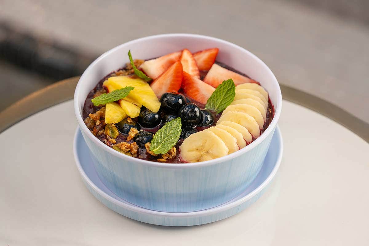 A bowl of acai topped with sliced banana, strawberries, blueberries, and mint leaves, on a white surface.