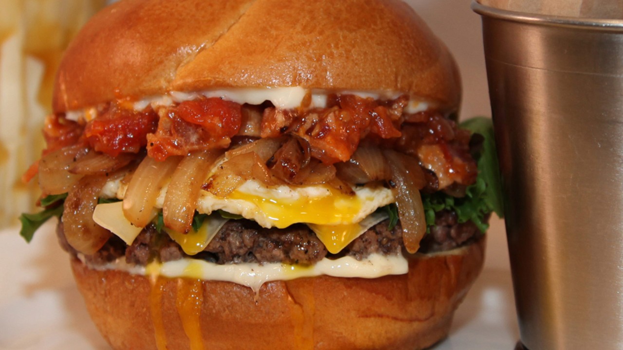 Close-up of a gourmet burger with melted cheese, caramelized onions, a fried egg, lettuce, and tomato relish in a brioche bun.