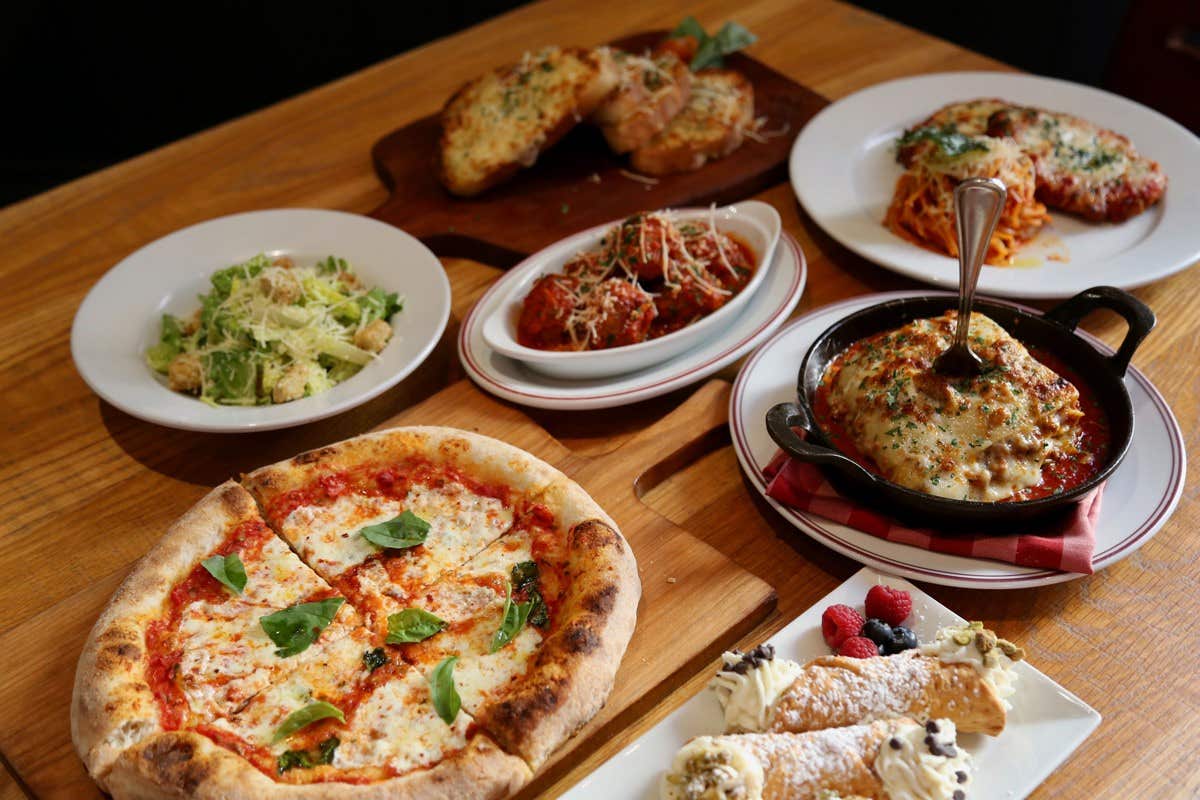 A table with various Italian dishes: pizza, salad, bread, meatballs, lasagna, spaghetti, and cannoli desserts.