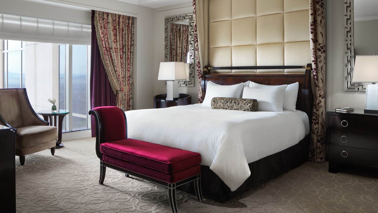 Elegant hotel room with a large bed, red bench, chair, nightstands, lamps, and a window with a cityscape view.