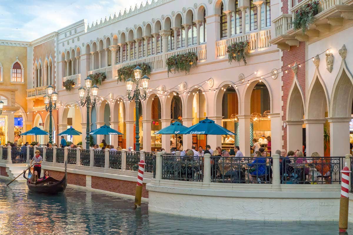 A gondola rides along a canal inside a shopping mall with outdoor cafes, blue umbrellas, and people dining.