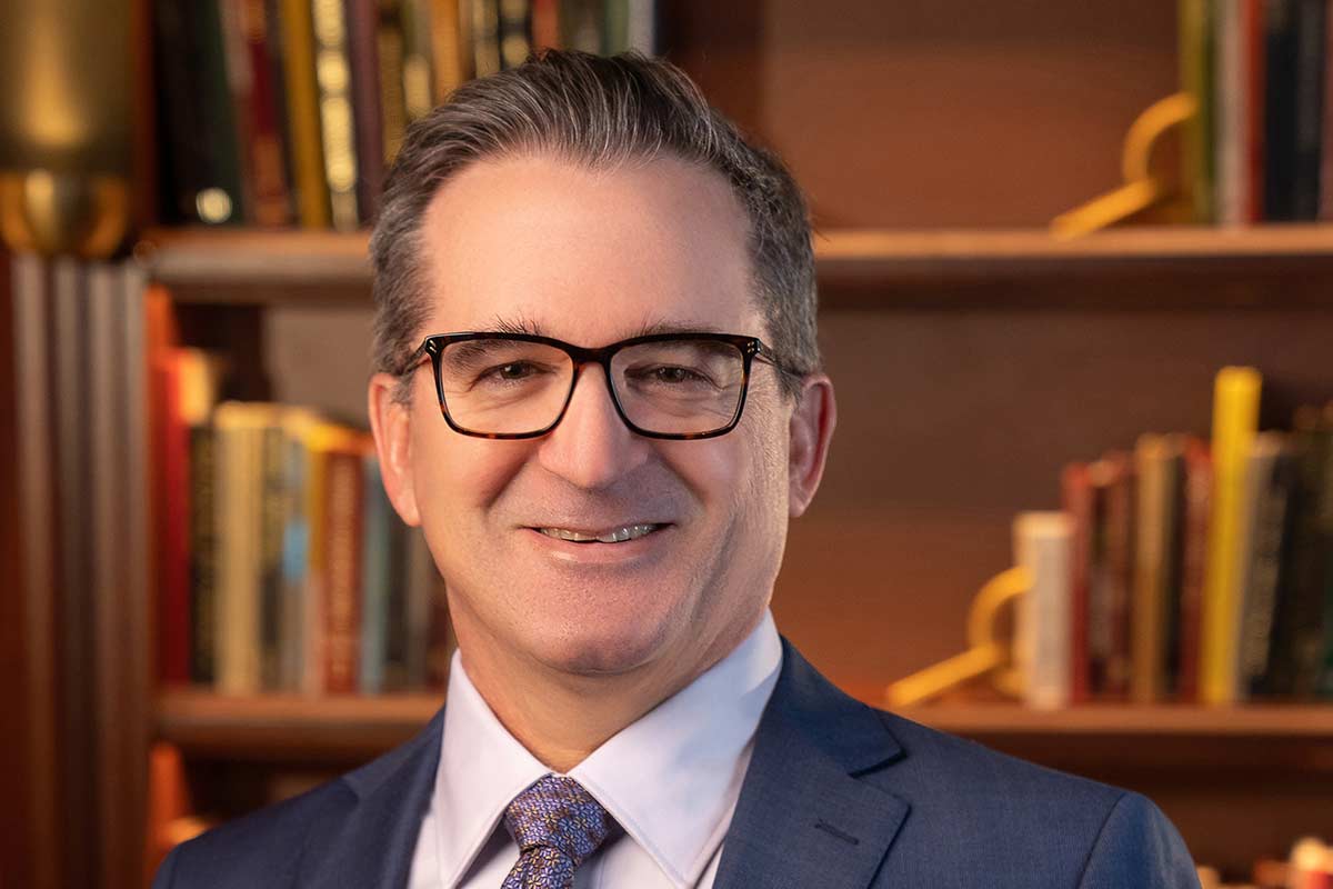 Smiling man in glasses and a suit stands in front of a bookshelf.