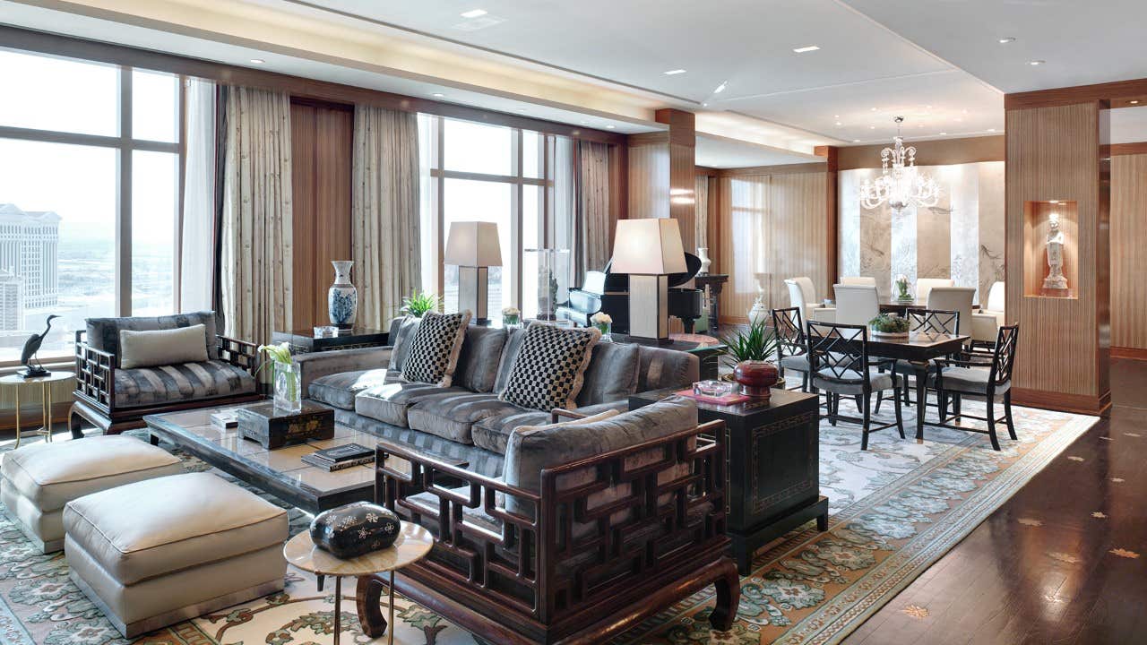 Luxurious living room with elegant furniture, large windows, and a dining area with a chandelier in the background.