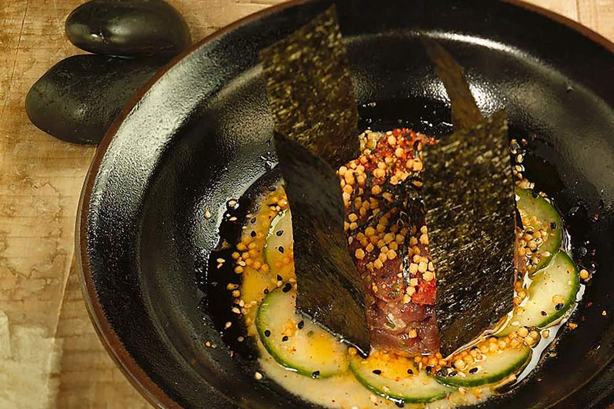 A bowl of sashimi topped with seaweed, cucumber slices, sesame seeds, and seasoning in a decorative black dish.