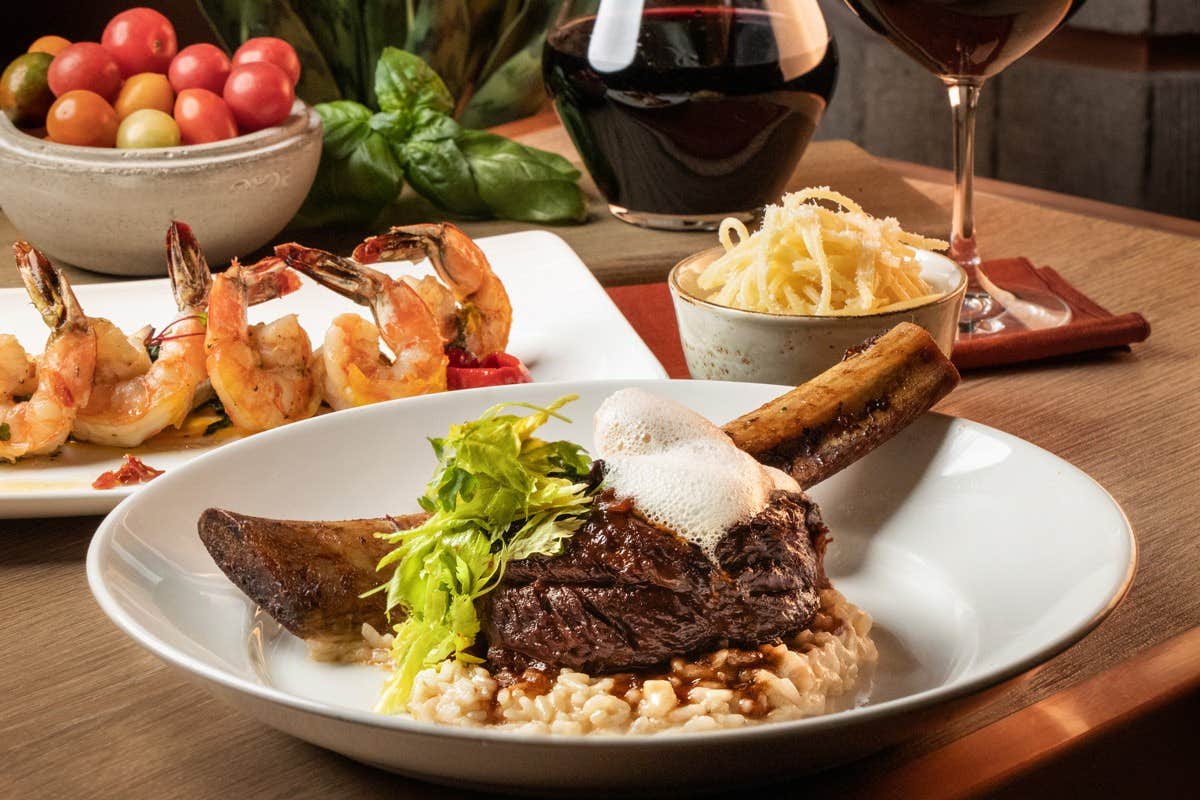 A gourmet meal with braised beef on risotto, grilled shrimp, cherry tomatoes, pasta, and red wine on a wooden table.