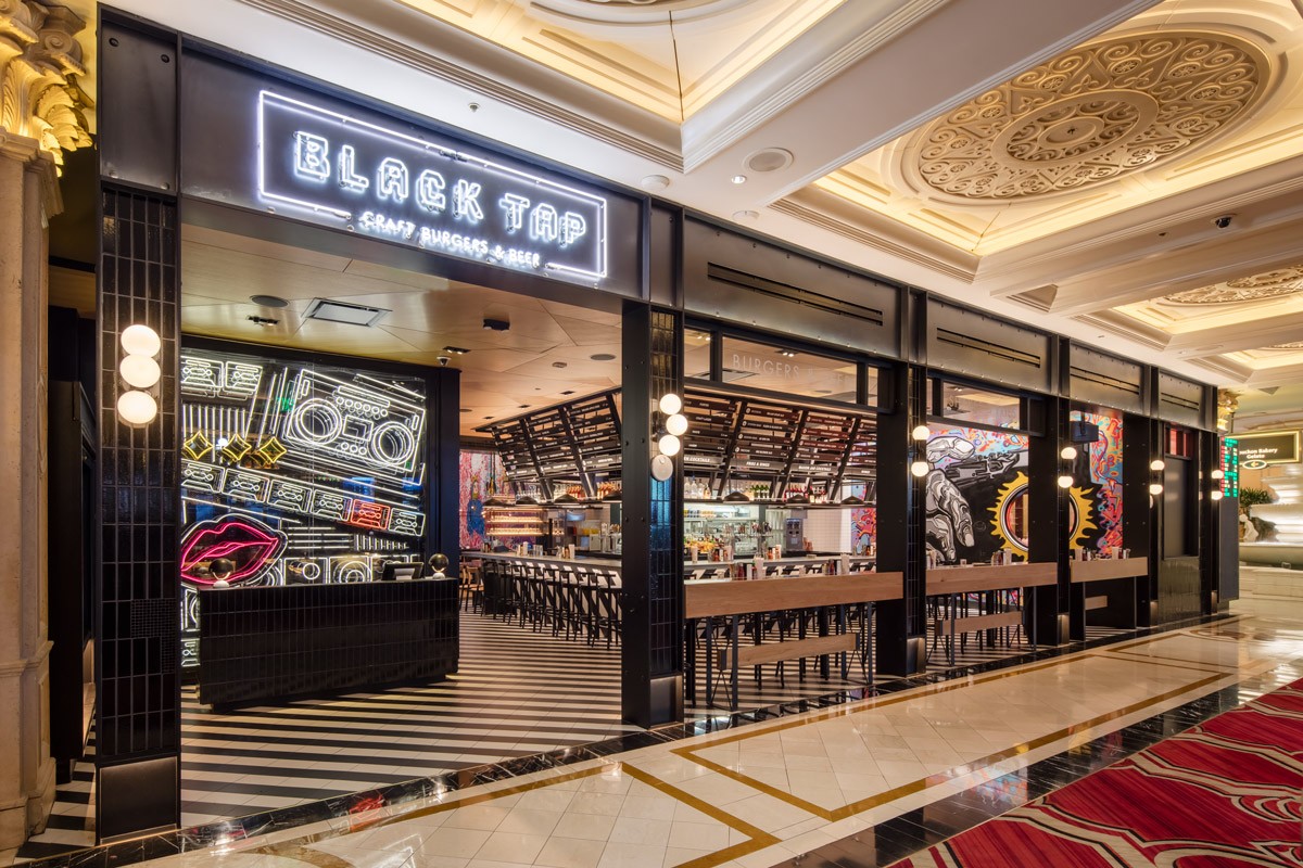 A stylish restaurant with neon signs and modern decor, featuring "Black Tap Craft Burgers & Beer" signage and artistic murals.